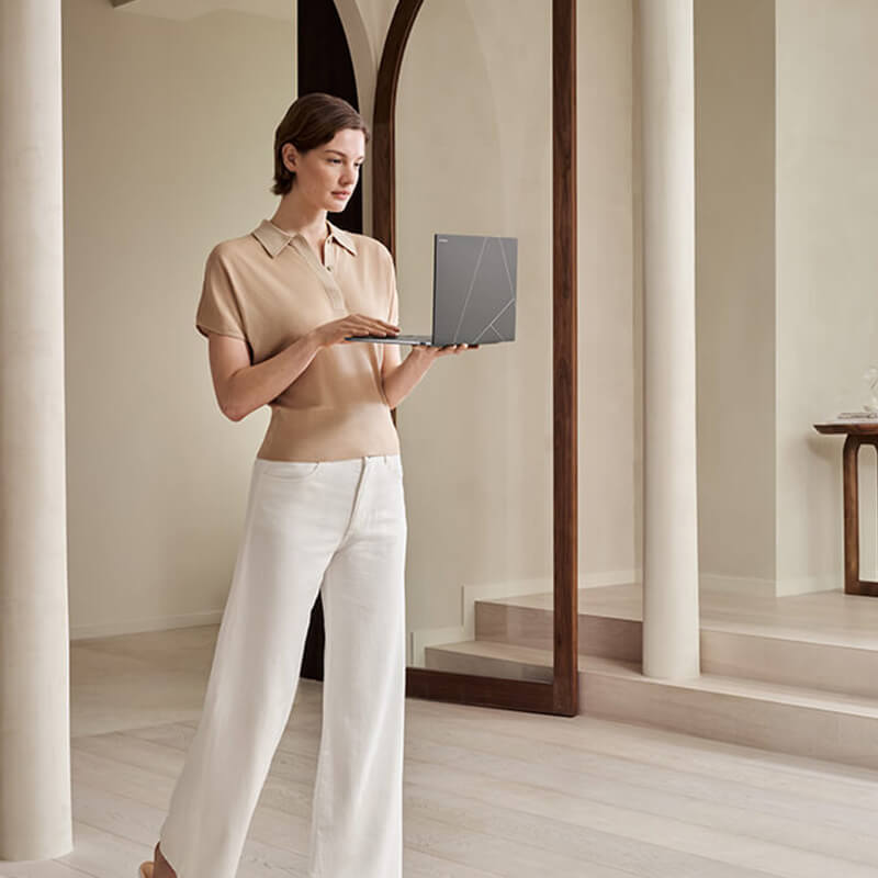 A young woman standing in a light-filled living room holding an open Zenbook S 14 laptop in her hands