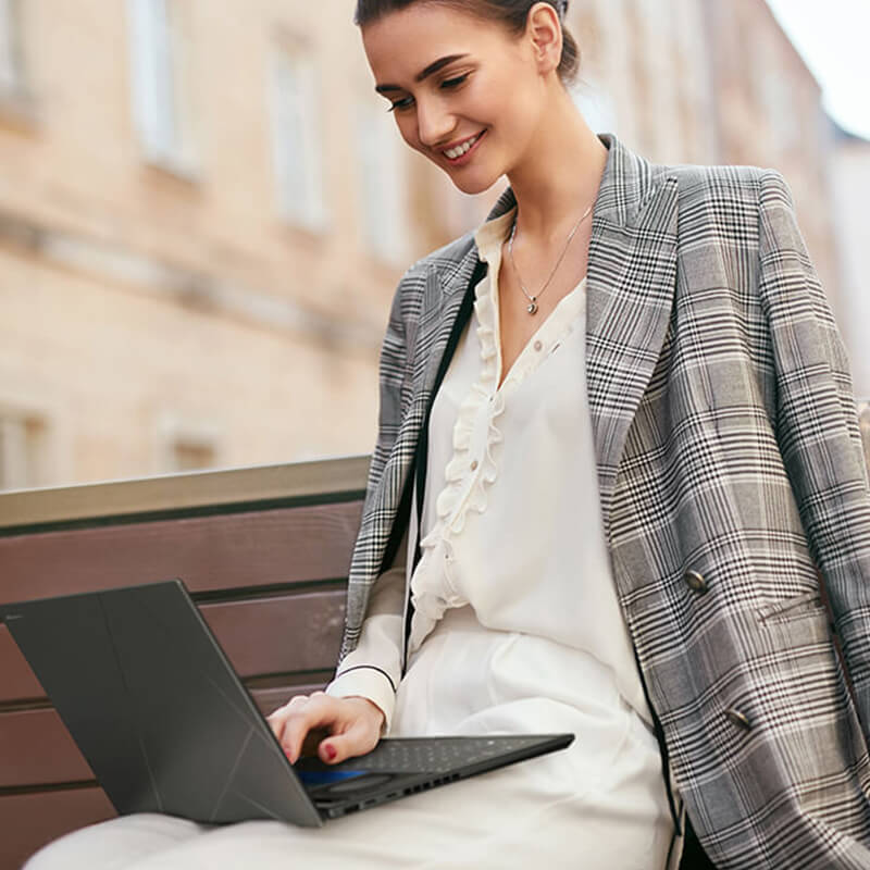 a young woman using ASUS Zenbook DUO laptop on her lap while sitting on a bench outdoors