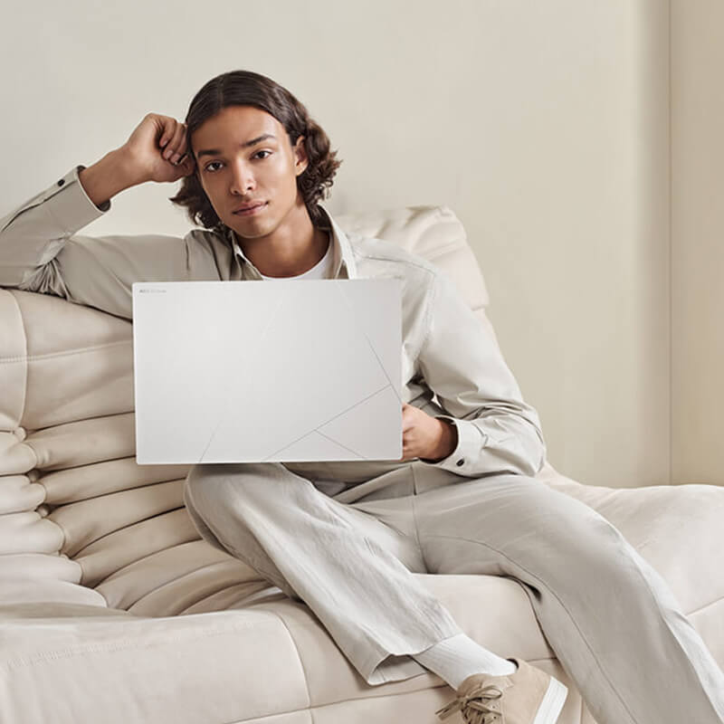 a young man sitting on a white sofa with a Zenbook S 16 laptop in Scandinavian White color on his lap