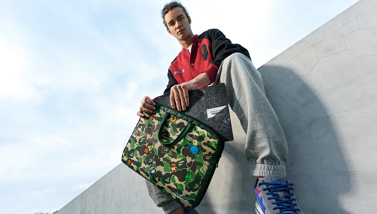 a young man sitting on a wall, dressed in a BAPE outfit, taking the ASUS Vivobook S 15 OLED BAPE Edition out of limited-edition laptop bag