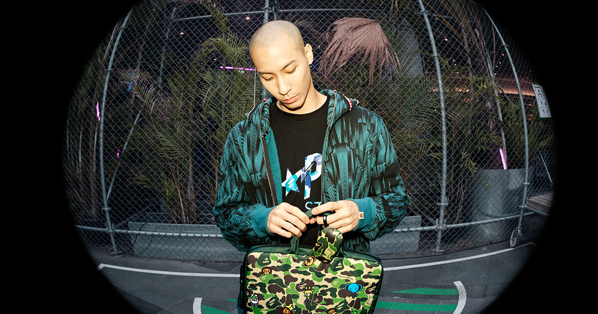 a young man dressed in A BATHING APE-branded hoodie and tshirt standing on an outdoor basketball court holding the limited edition ASUS and BAPE collab laptop bad and mouse with the green camo design