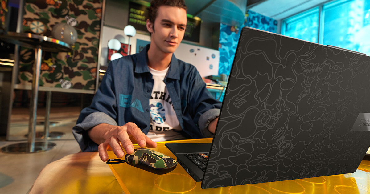 a young man dressed in BAPE clothes using a black ASUS Vivobook S 15 OLED BAPE Edition laptop and ASUS Marshmallow mouse BAPE edition, with the reflective camo pattern on matte black background on the laptop lid