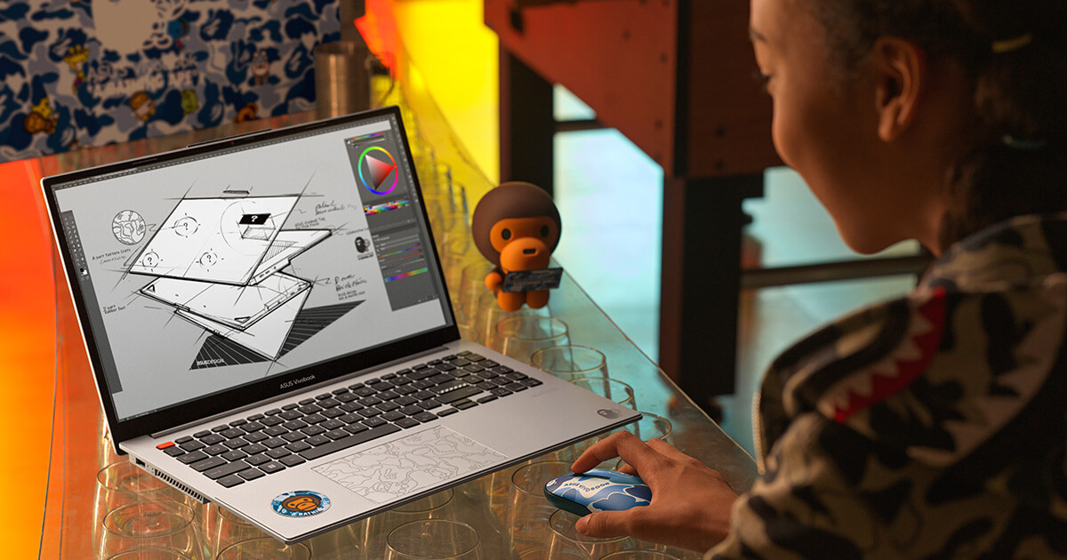 a young woman wearing a BAPE hoodie with signature BAPE camo pattern using a silver ASUS Vivobook S 15 OLED BAPE Edition laptop and blue camo design ASUS marshmallow BAPE Edition mouse for graphic design with a BABY MILO figurine placed next to the laptop