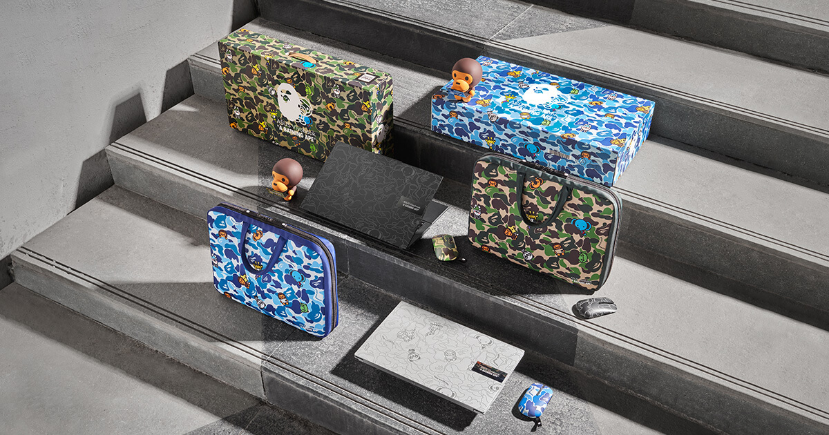 ASUS Vivobook S 15 OLED BAPE limited edition laptop bundles in green and blue colorways sitting on concrete stairs, with all the accessories lied out next to the black and silver laptops