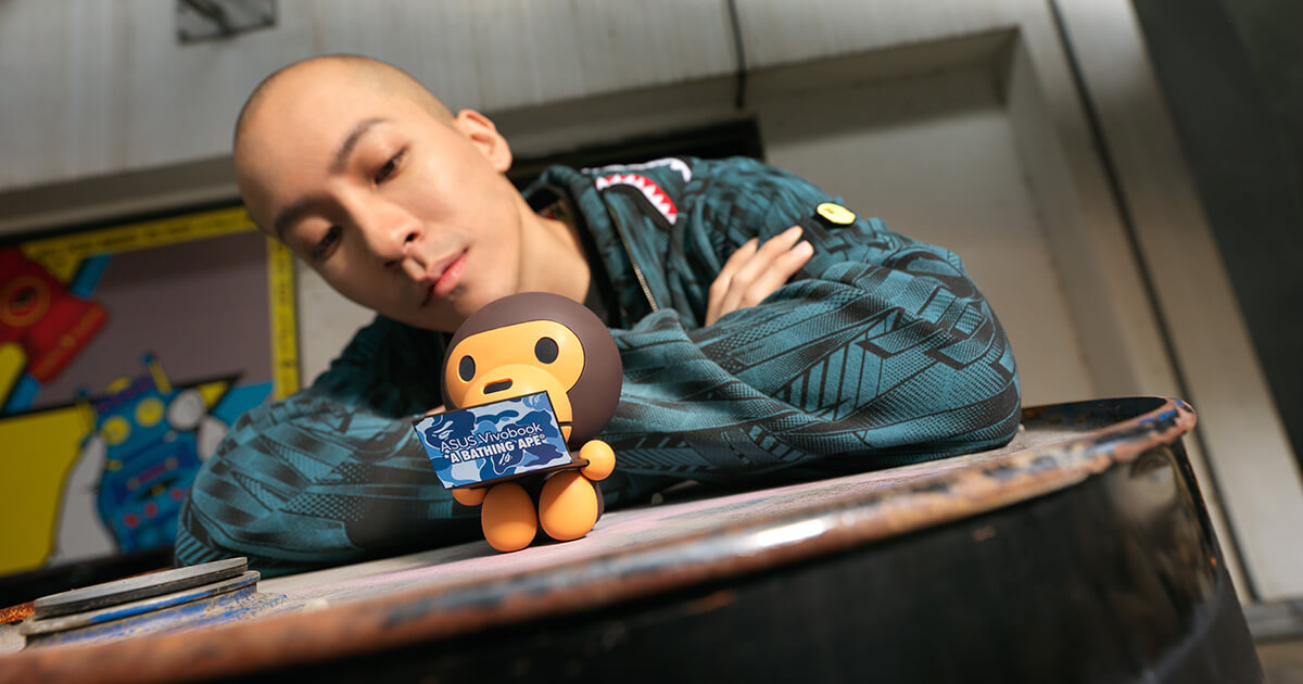 a BABY MILO figurine holding a miniature ASUS Vivobook laptop with a young man dressed in BAPE clothes in the background