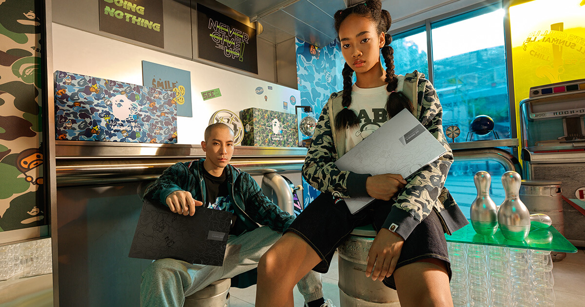 two young people sitting in a modern, streetwear-inspired room while holding the limited edition ASUS Vivobook X BAPE collab laptop in black and silver colors