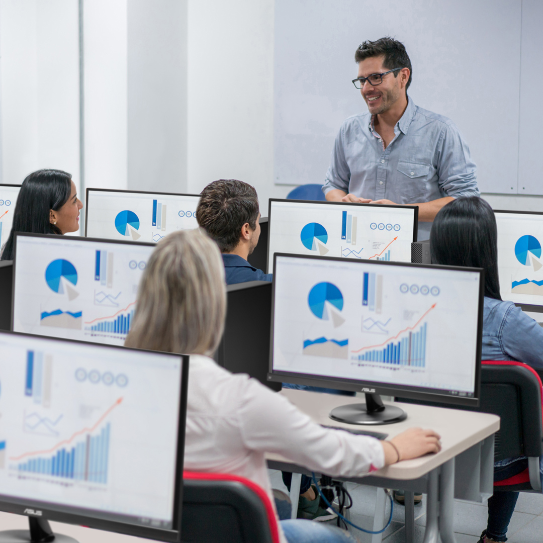 A teacher is teaching in a computer lab, standing in front of the classroom. There are several students sitting in the classroom with ASUS ExpertCenter desktops and monitors on the desk