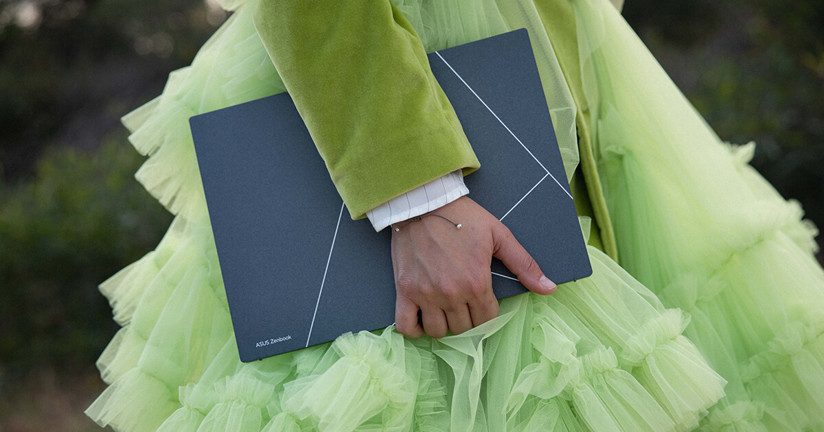 A woman in a green dress is holding Zenbook S 13 OLED. The image is captured from the side and focuses on the woman’s hand gripping the laptop.