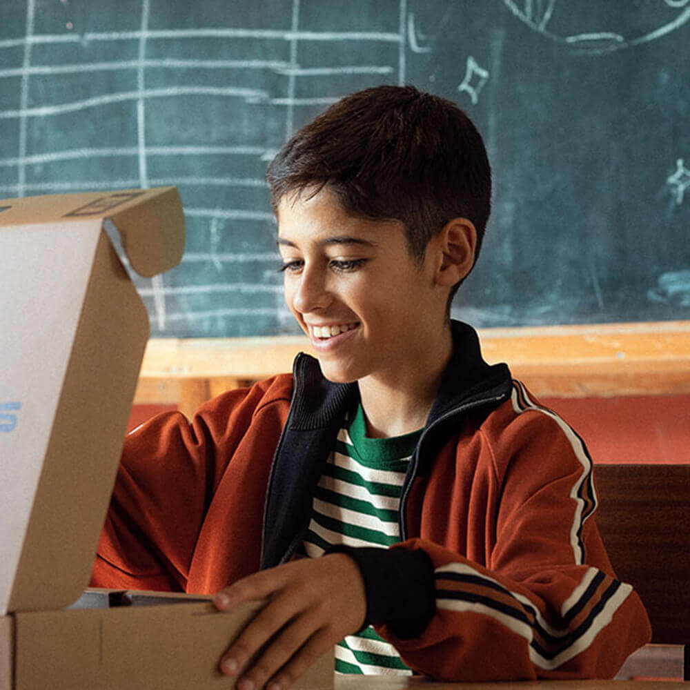 Uśmiechnięty uczeń otwierający pudełko z laptopem ASUS, siedząc przed tablicą w klasie