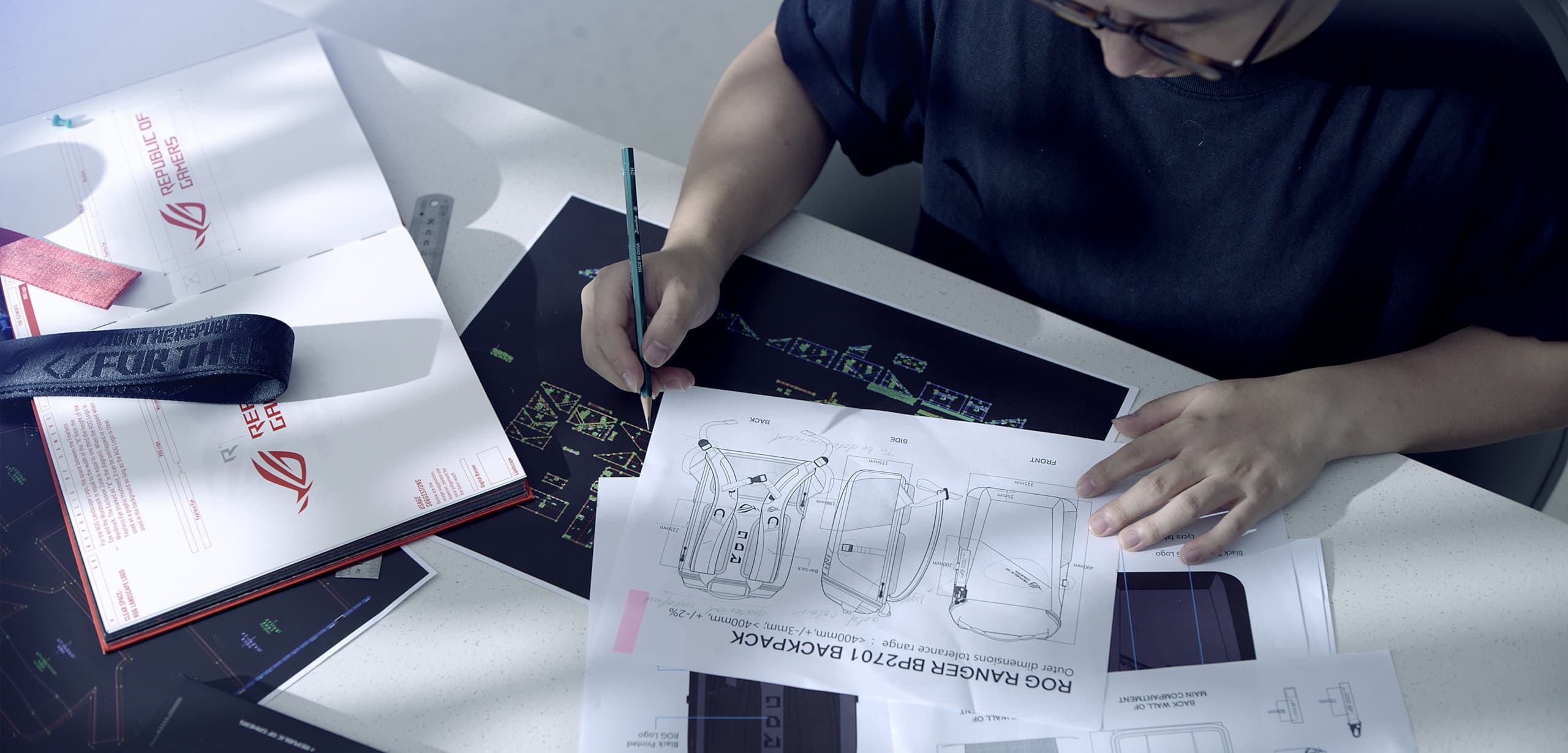 Man sketching on a table, with different designs and pictures of the ROG Ranger Backpack shown on the table arranged in front of him.
