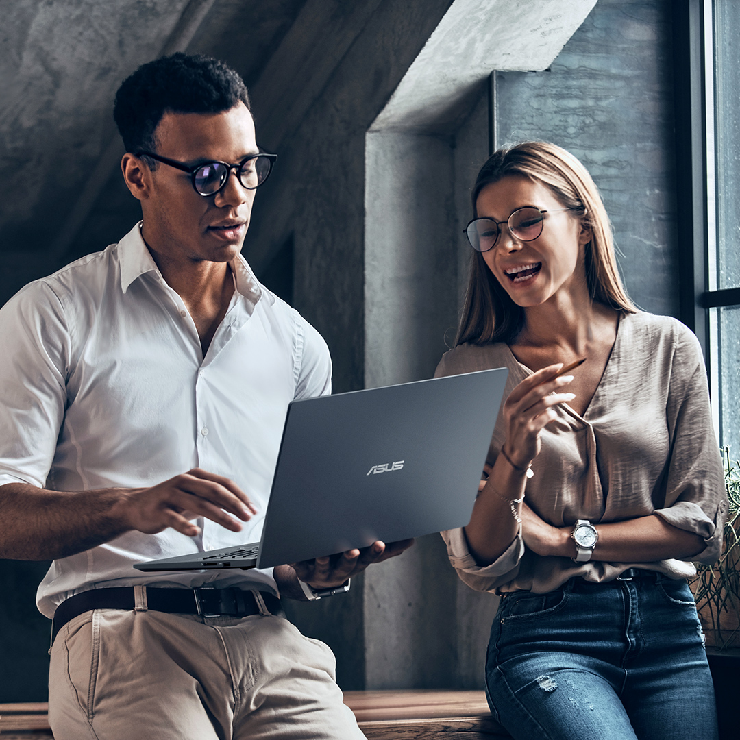 Dwóch pracowników dyskutuje radośnie, trzymając w ręku laptopa ASUS..