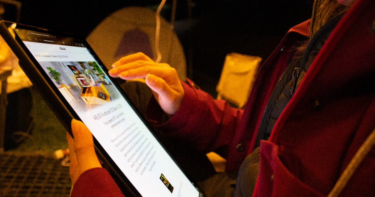 A woman holding ASUS Vivobook 13 Slate OLED convertible laptop in tablet mode in one hand, with a camping chair and tent in the background