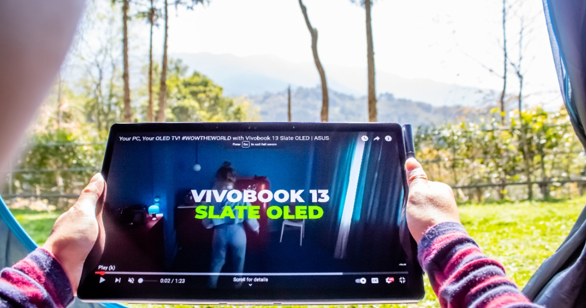 a woman watching a video on ASUS Vivobook 13 Slate OLED convertible laptop, holding it in two hands in horizontal table mode while sitting in a tent, during sunny day, with grass, trees, and mountains in the background