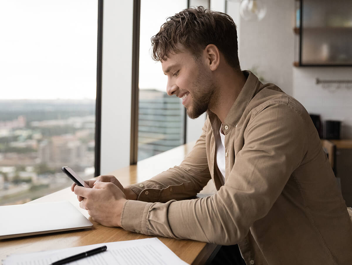 A man sitting in a cafe by a window, smiling and looking at his smartphone.