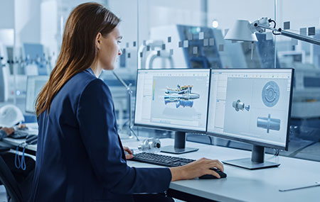 A lady in office sitting in front of a desk with 2 monitor displays, a keyboard, and a mouse. The screens show 3D rendering of vehicle parts