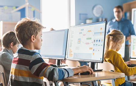Three elementary school students in the classroom with one teacher up front. Each students sit in front of a desk monitor working on their assignments