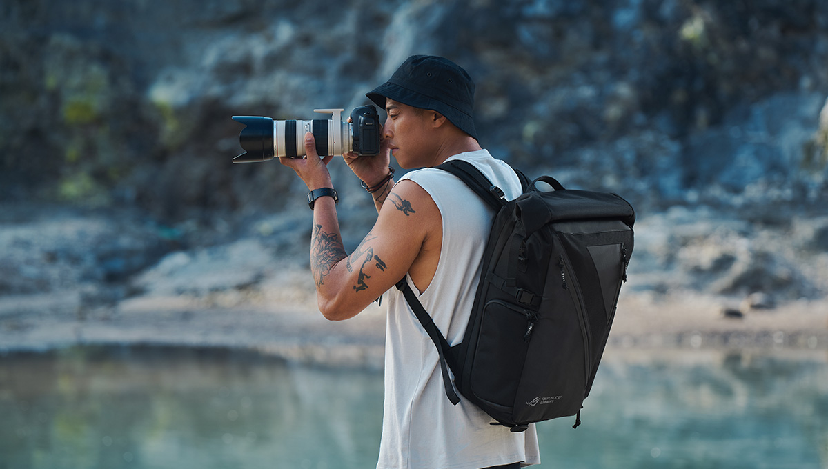 a young photographer taking a photo while outdoors with an ROG Archer Backpack 17 camera backpack on his back