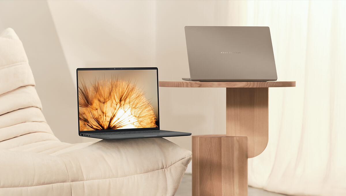 Two ASUS Zenbook 14 laptops sit open in a modern-looking room, one on the edge of a sofa and one on a wooden table beside the sofa.