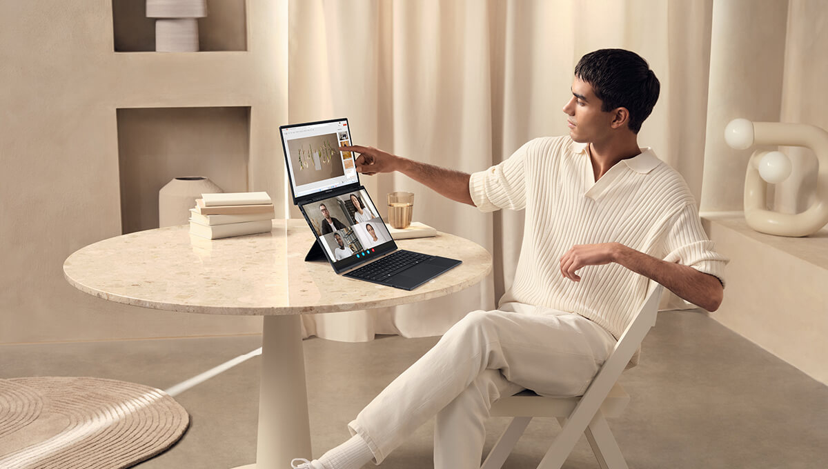 A young man dressed in light-colored clothes in a bright, modern room touches the upper screen of an open ASUS Zenbook DUO laptop in Dual Screen mode