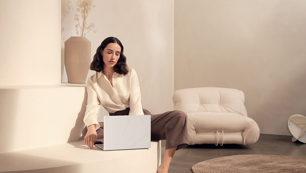 A young woman sits on a sofa, looking at an open ASUS Zenbook S16 laptop.