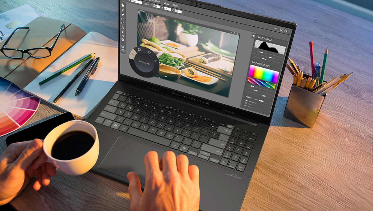 A person uses the ASUS DialPad on an ASUS Vivobook Pro 15 laptop to edit an image with one hand while holding a cup of coffee in the other.