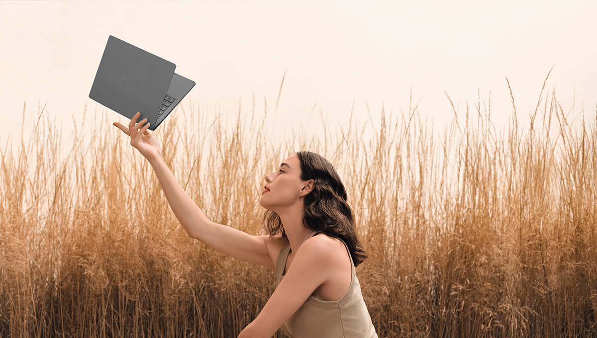 A young woman holds an ASUS Zenbook 14 laptop on top of her held-up hand while squatting in front of a grain field.