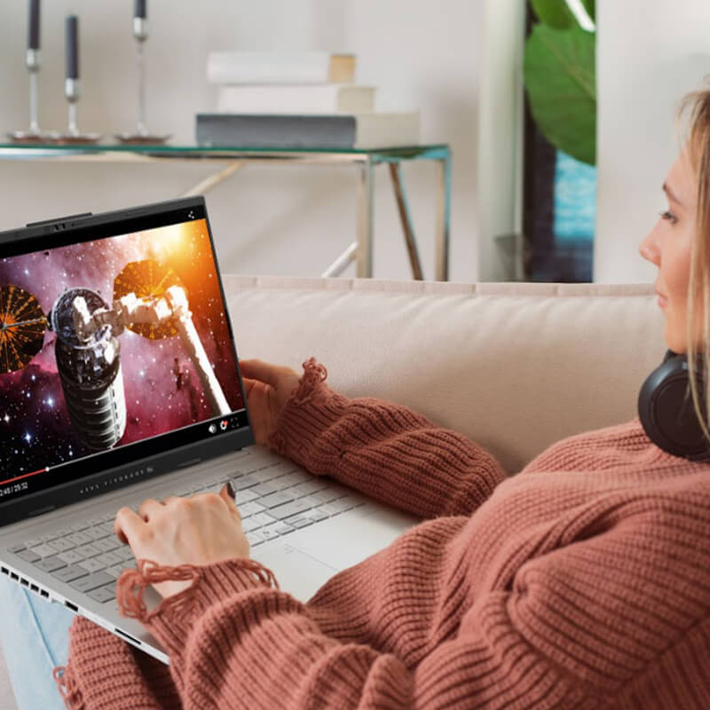 A young woman sitting on a pink sofa with an ASUS laptop on her lap, watching an online video