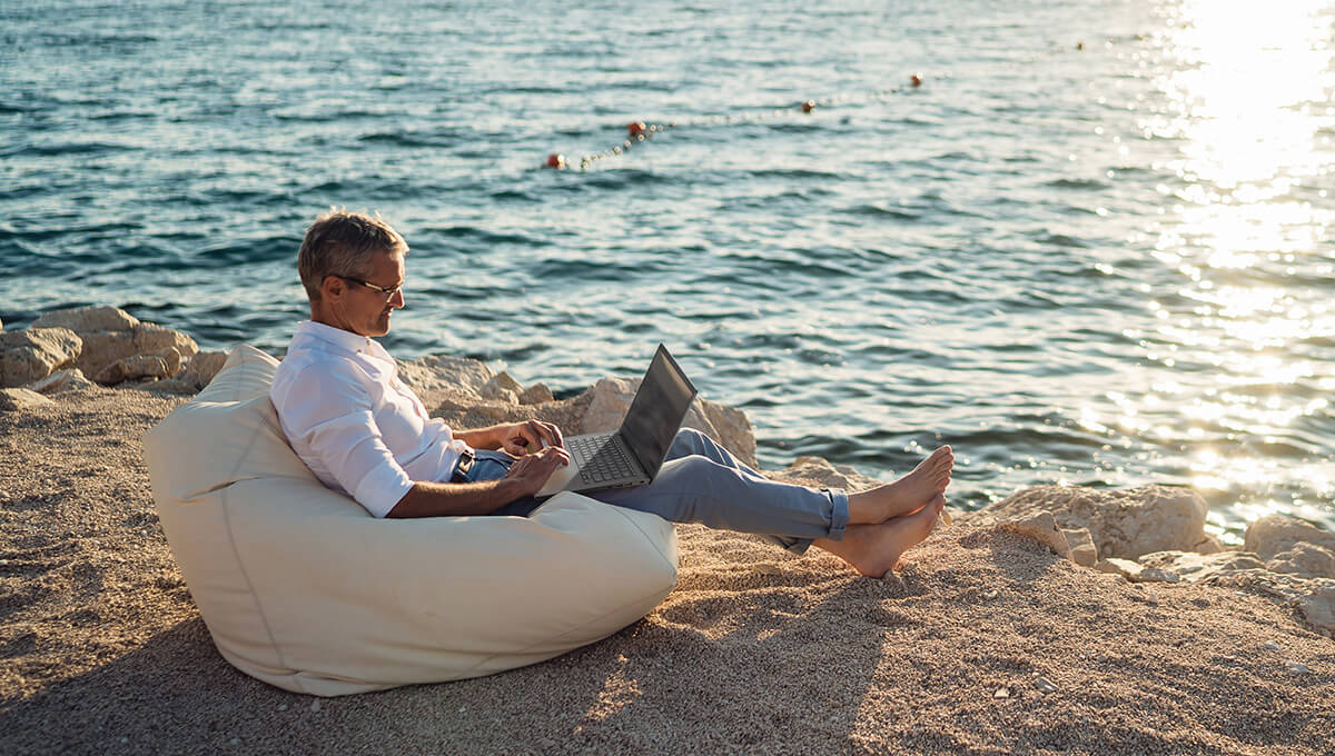 a middle-aged man sitting on a beach on a bean bag with an ASUS Vivobook laptop on his lap