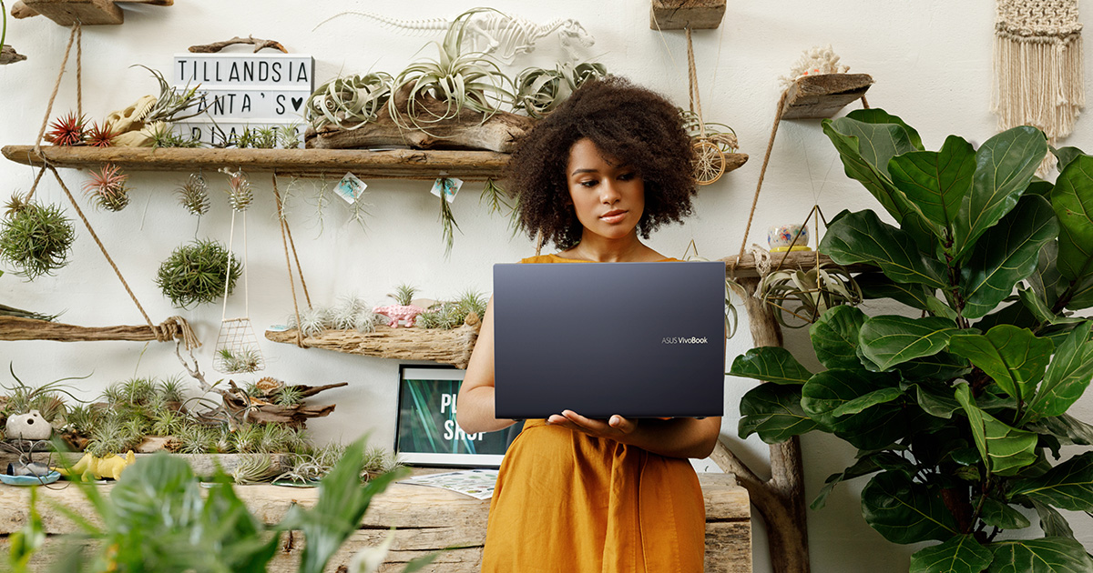 a young woman holding an ASUS Vivobook 15 OLED laptop in one hand and typing with another hand while standing in a plant shop