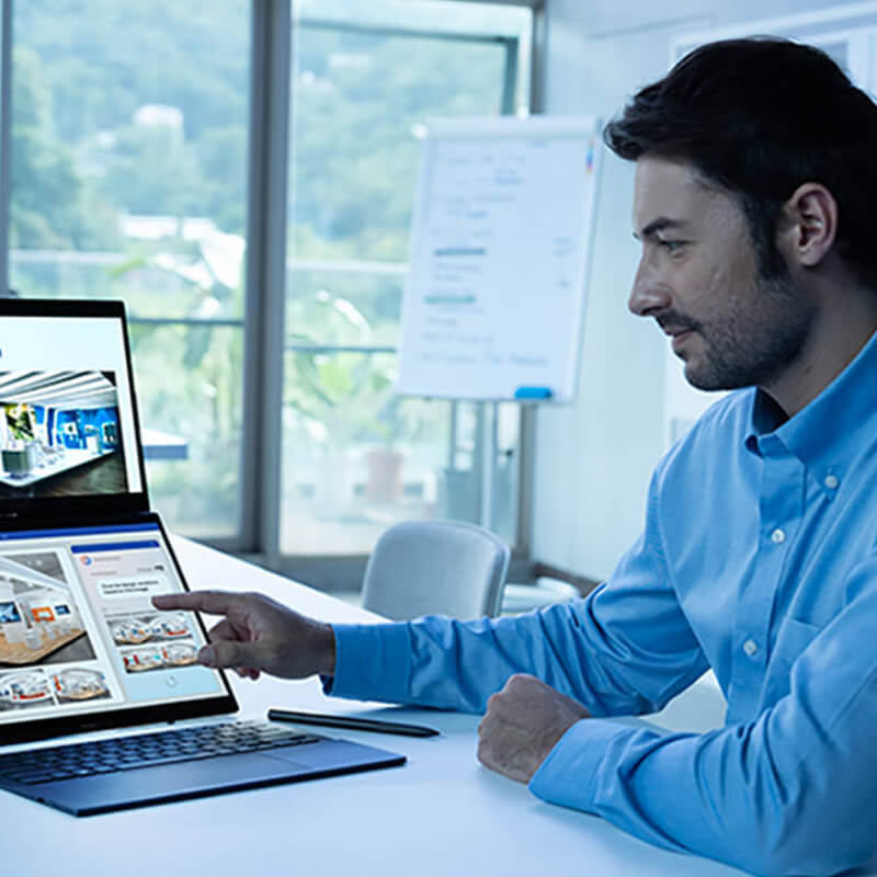 Un hombre con camisa azul apunta a la pantalla del Zenbook DUO en una sala de reuniones blanca. La pantalla de la parte superior muestra su proyecto principal, mientras que la de la parte inferior muestra ventanas divididas.