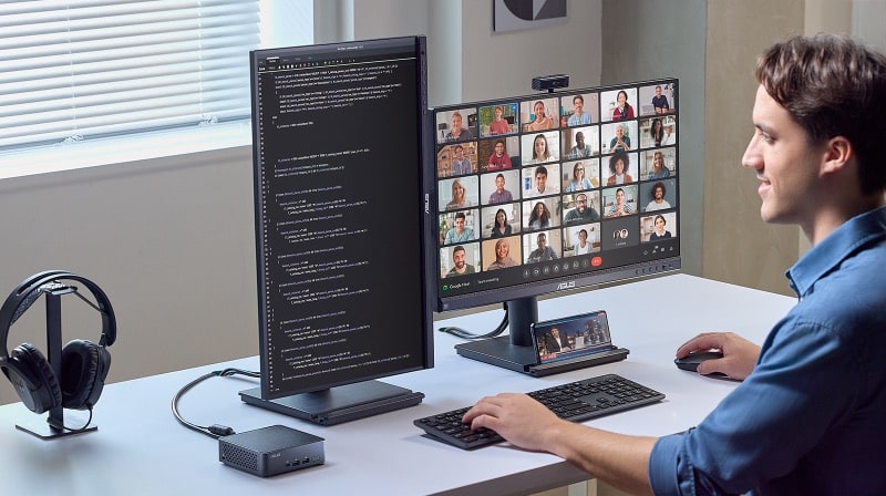 A person at a desk uses dual monitors and a best mini PC — one screen shows code, the other a video call with multiple people.