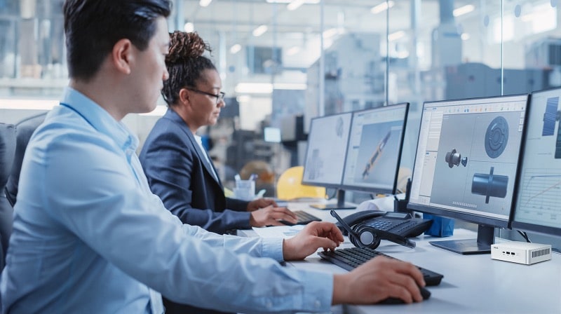 Two engineers work on CAD designs in a modern office, using large screens powered by a silver best mini PC.
