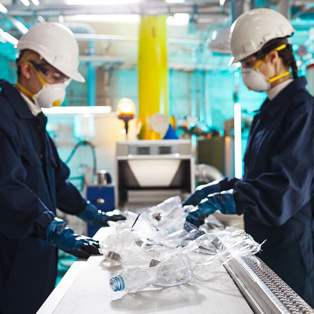 A man and a woman in blue jumpsuits, masks, and hard hats are work at a conveyer belt, sorting plastic bottles.