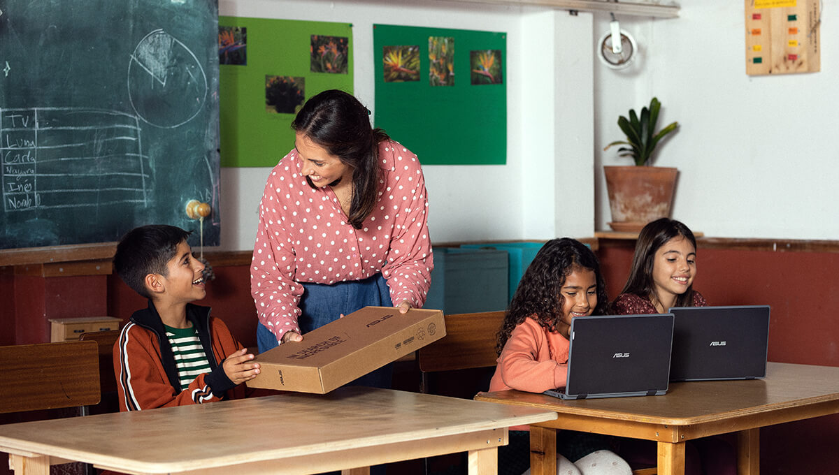 A female teacher in a classroom hands a young boy student a box that contains a laptop, and two young girl students smile as they use their ASUS laptops on a nearby desk.