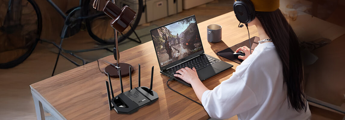 Dziewczyna w żółtej czapce i słuchawkach siedzi przy drewnianym biurku i gra na laptopie. W pobliżu znajduje się router i lampa biurkowa.