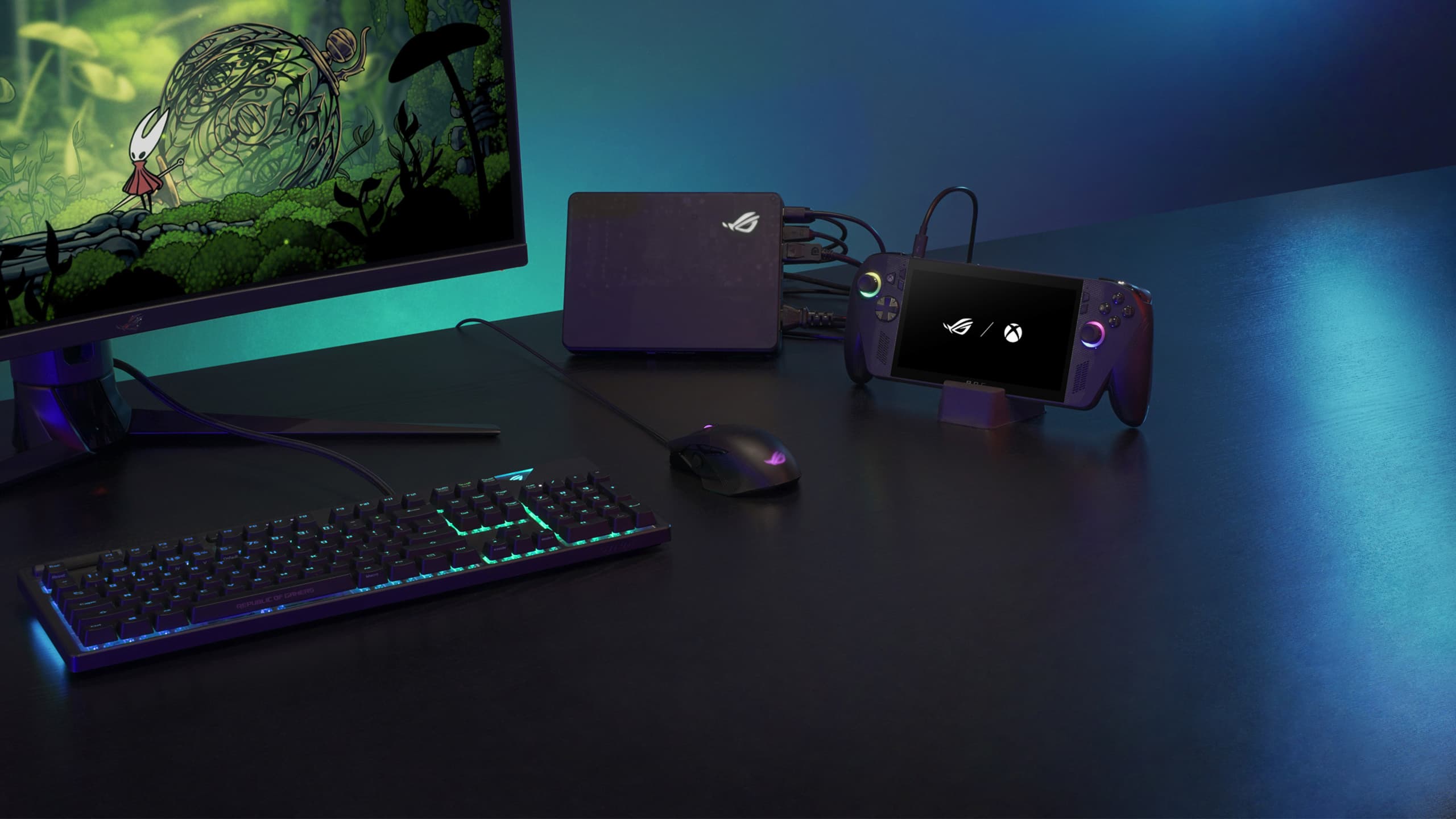 An ROG Xbox Ally X in a stand, headphones, a mouse, a keyboard, and a monitor arranged on a desk in a darkly lit room.