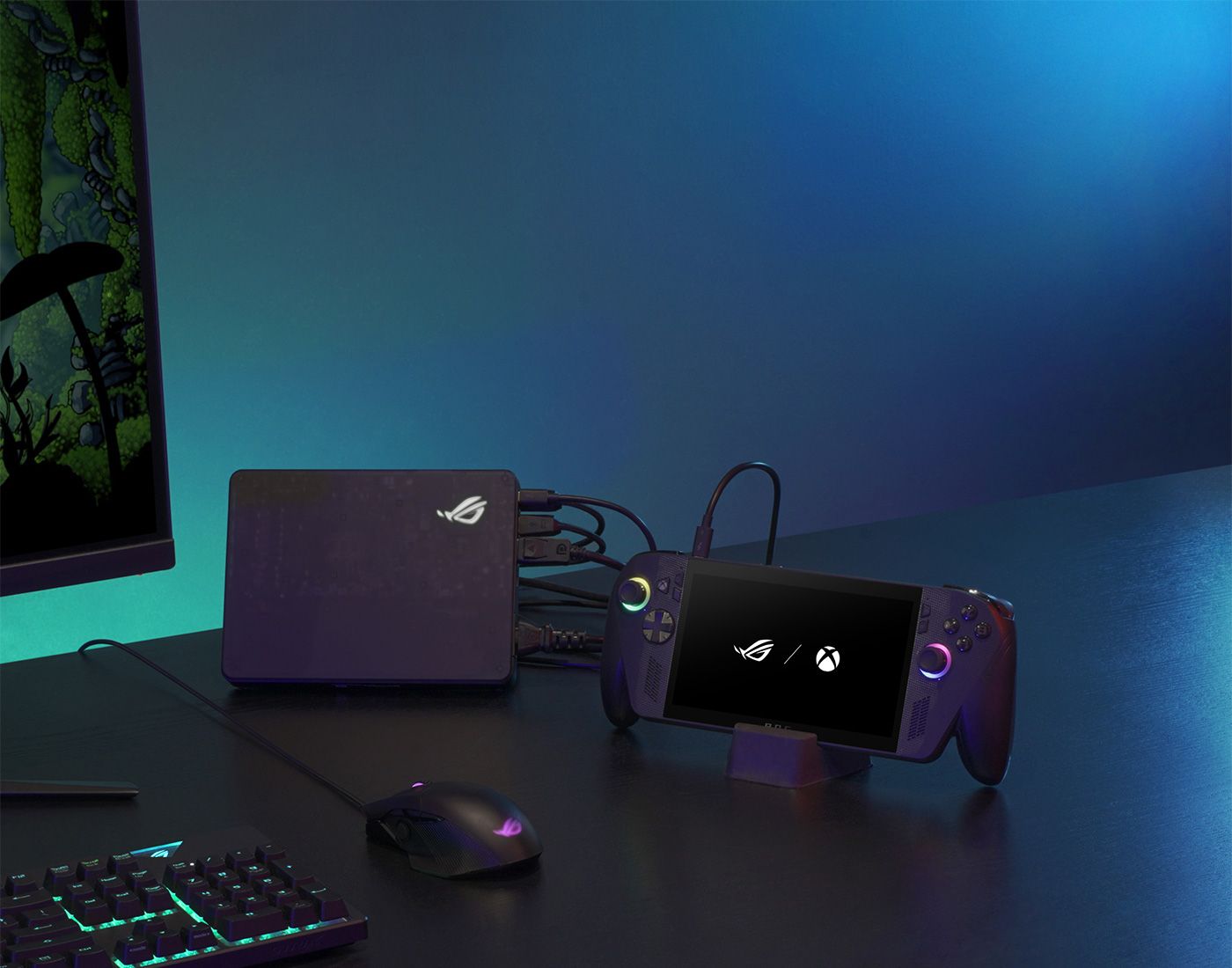 An ROG Xbox Ally X in a stand, headphones, a mouse, a keyboard, and a monitor arranged on a desk in a darkly lit room.