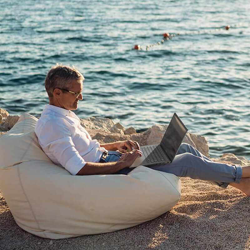 a middle-aged man sitting on a beach on a bean bag with an ASUS Vivobook laptop on his lap