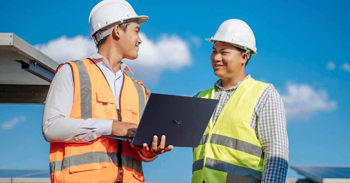 Two men in neon working vests and hard hats talk, the man on the left holds an ASUS laptop.