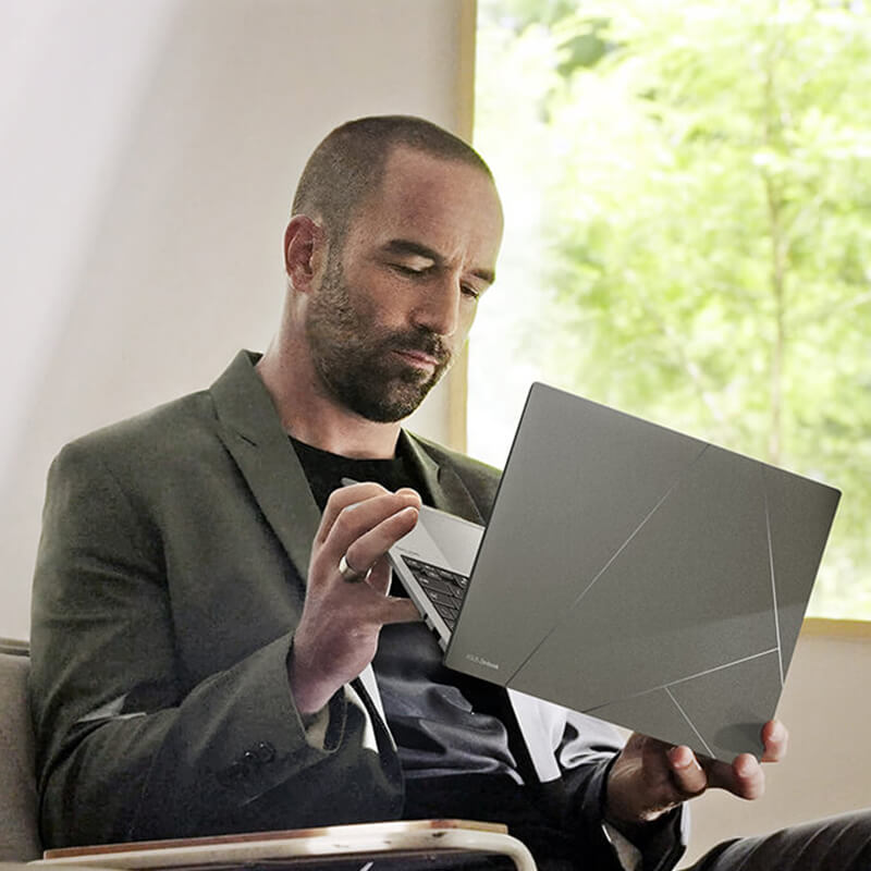 A man in smart casual attire is holding Zenbook S 13 OLED basalt grey version and inspecting its appearance. The background is beige with greenery on the right.