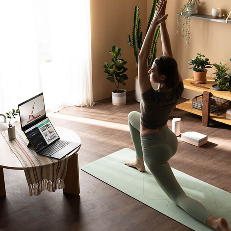 A woman on a light green yoga mat is doing yoga on the right. She is looking at a dual screen laptop propped on the round table on her left. There are two screens on the laptop. The display on top is showing the woman’s yoga tutorial while the one on the bottom is in split screen showing different information.