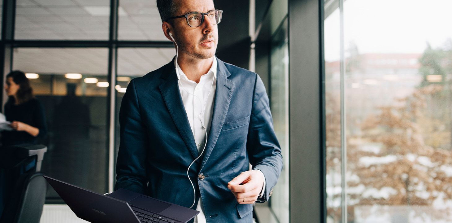 An executive wearing earphones while holding the ASUS ExpertBook laptop in his hand and standing by the window in the office.