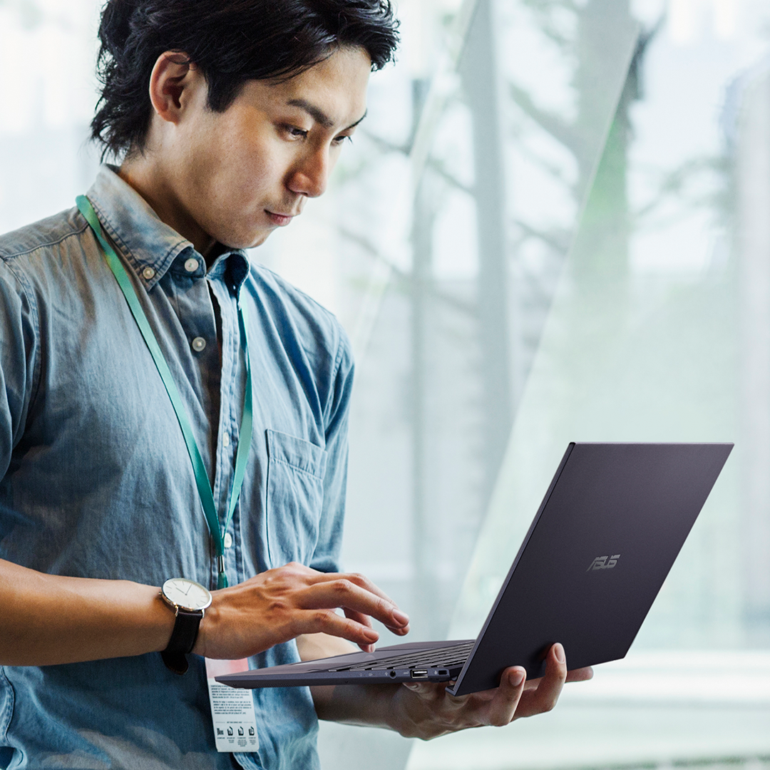 Un uomo d'affari in piedi accanto alla finestra dell'ufficio utilizza un computer portatile ASUS ExpertBook.




