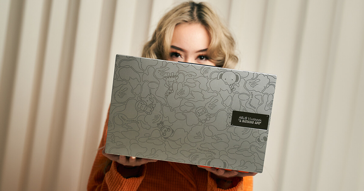 A woman is holding a silver ASUS Vivobook BAPE® Edition with half of her face obscured by it. The photo light etched patterns are clearly visible as the subject of the image.