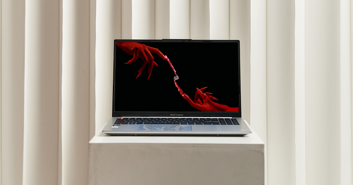 A silver laptop is propped on a white display stand. An image of two red hands coming from two diagonal sides of the picture with long nails holding a chip at the center is shown on the laptop screen.