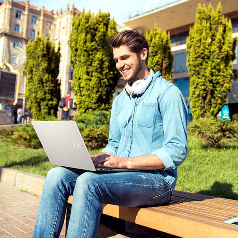 a college student sitting in front of a university building with an open ASUS Vivobook Pro 15 OLED laptop on his laps
