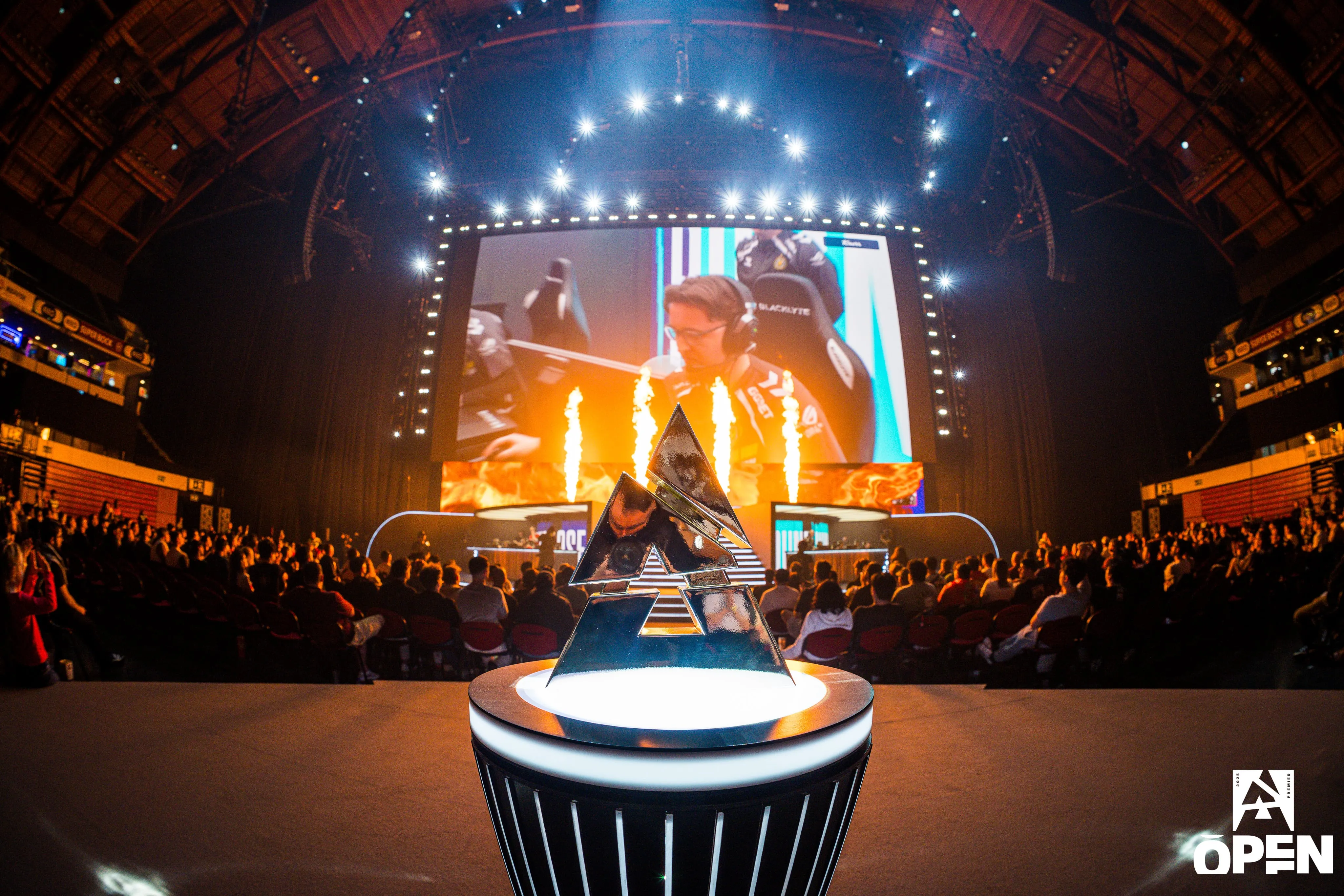 BLAST Premier esports event stage with trophy in the foreground, large screen showing a player, and audience watching amid bright stage lights and flame effects.