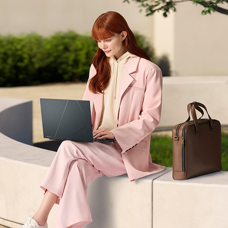 Девушка сидит на ограде с ноутбуком ASUS Zenbook 14X OLED на коленях. Рядом с ней стоит коричневый портфель, а на заднем фоне видно дерево