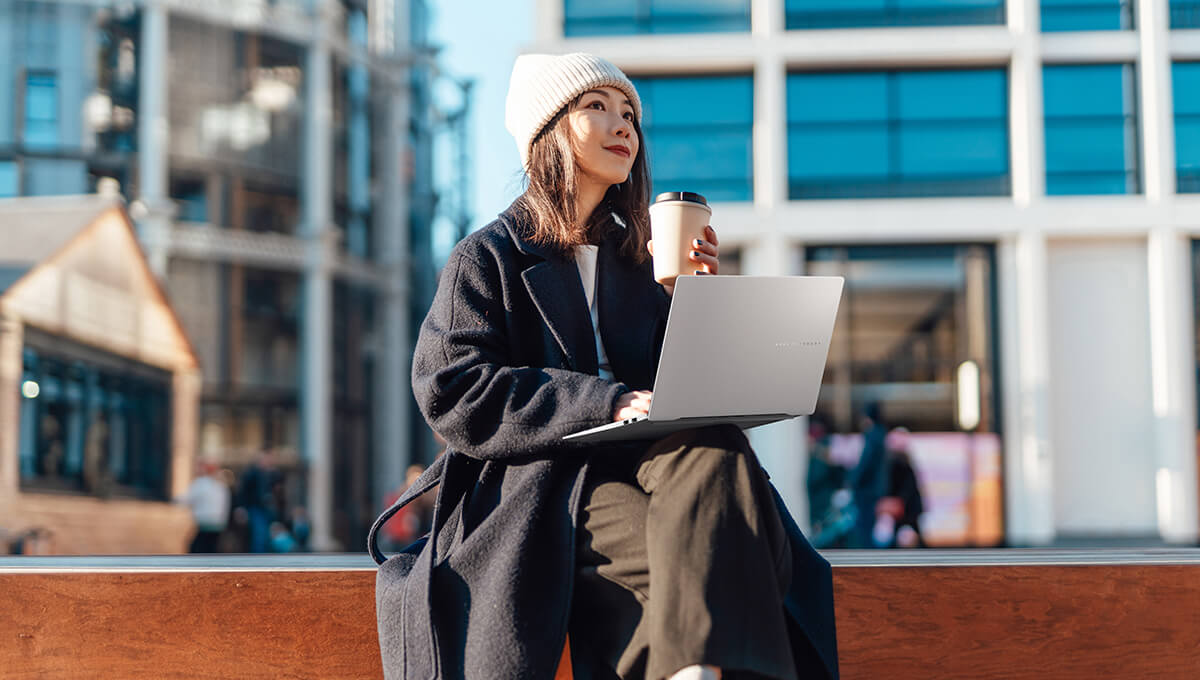 A young woman sits on a bench outdoor with a cup of coffee and an ASUS Vivobook S 14 OLED laptop on her lap