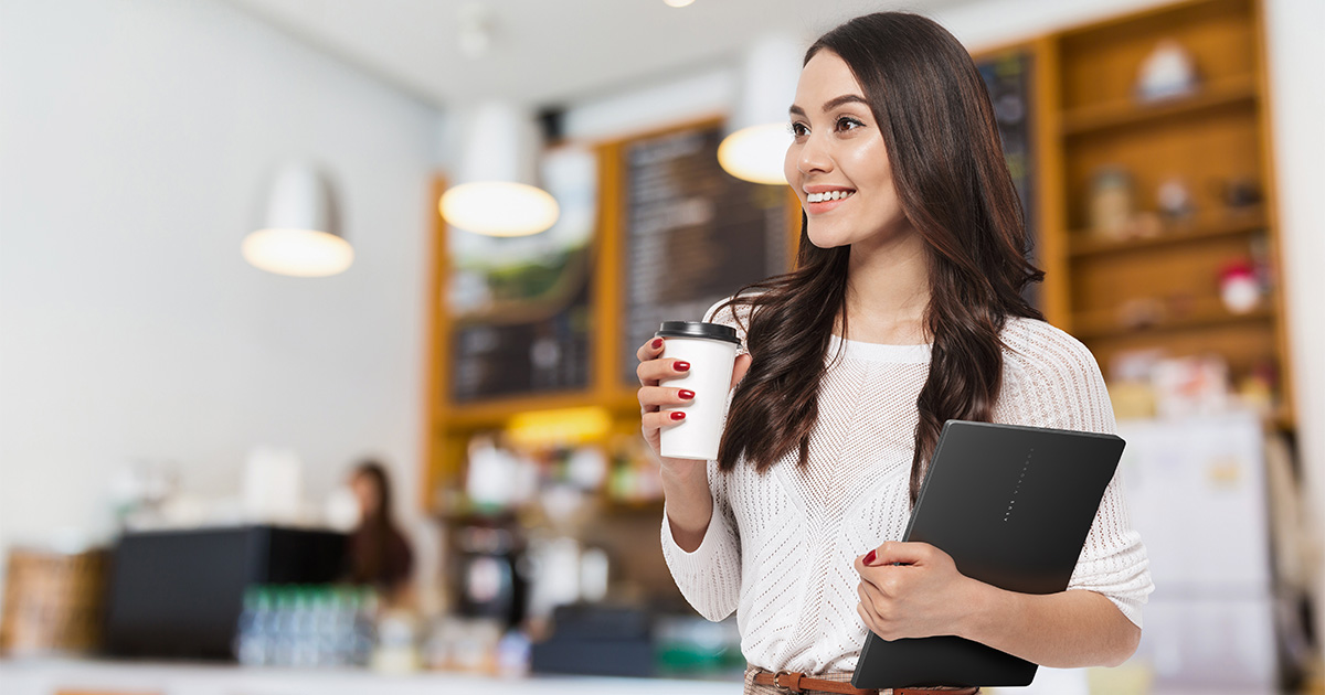 A smiling woman is holding ASUS Vivobook S 14 OLED in one hand and a cup of coffee in the other.